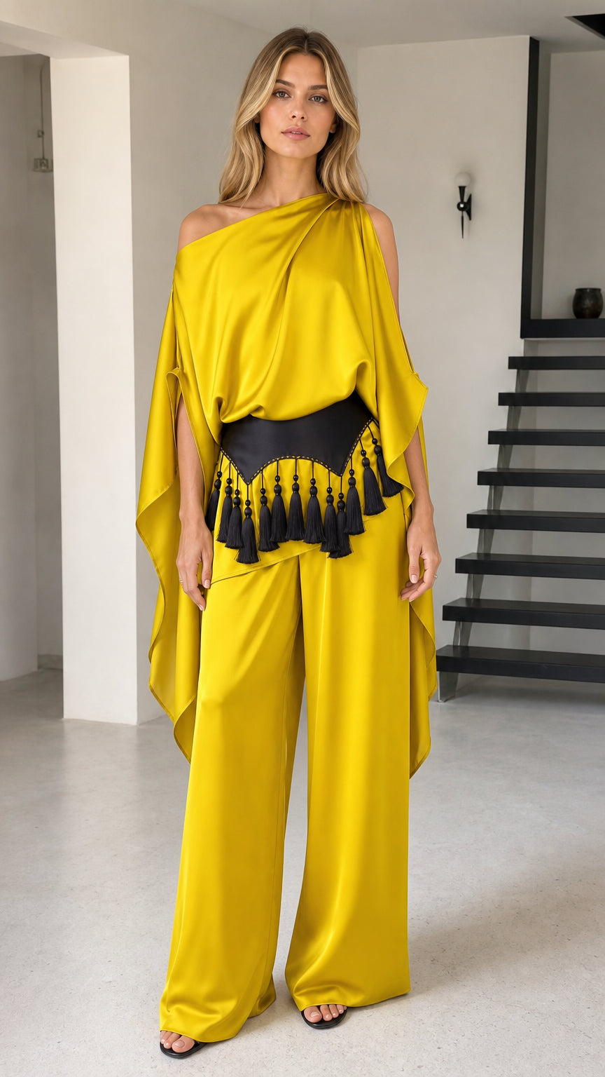 Front view of model wearing yellow one-shoulder cape top and wide-leg pants set with black tassel belt